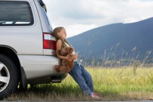 A little girl leaning against a car on the side of the road holding a teddy bear.